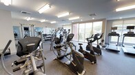 This fitness center is a thoughtfully designed workout space housed on a single floor. The room is illuminated by a combination of recessed lighting and large overhead fluorescent fixtures, ensuring bright, even light throughout the space. A row of expansive windows along one wall floods the room with natural light while providing views of the surrounding outdoor greenery, creating an open and energizing atmosphere.

The cardio section is well-stocked with multiple elliptical trainers, stationary bikes, and additional aerobic equipment, all arranged to maximize usable floor space. A full-length mirror runs along one wall, enhancing the sense of depth and allowing users to monitor their form during workouts. Adjacent to the mirror, a dedicated free weights area features a neatly organized dumbbell rack with a wide range of weight options.

The flooring throughout is a durable, textured rubber or sport-grade material suited for high-traffic gym use. The ceiling is finished in a clean, light tone with integrated ventilation to support air circulation. The overall layout is practical and uncluttered, offering a focused environment suitable for a range of fitness activities.