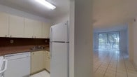 This single-floor space offers a straightforward and functional layout connecting a kitchen to an open living area. The kitchen is equipped with cream-colored cabinetry, a dark tile backsplash, a stainless steel sink, and a full-size top-freezer refrigerator. Overhead fluorescent lighting illuminates the workspace, and light tile flooring runs throughout. A structural column separates the kitchen from the adjacent living area, which continues the same tile flooring for a cohesive feel. The living area benefits from natural light streaming in through a large sliding glass door fitted with vertical blinds, offering a view of outdoor greenery. The overall color palette is neutral, featuring white walls and light flooring that contribute to a clean, open atmosphere. The single-floor design ensures easy navigation throughout the space, making it practical and accessible.