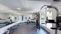 This fitness center offers a practical and well-organized space designed to support a comprehensive workout routine. The room features durable rubber flooring that provides both safety and comfort during exercise. Large floor-to-ceiling windows along one wall flood the space with natural light and offer views of lush outdoor greenery, creating an energizing atmosphere for workouts.

The facility is equipped with a selection of cardio and strength training machines, including an elliptical trainer, a cable resistance machine, and additional strength equipment. A rowing machine or bench is also visible along the side, further expanding the training options available. Full-length mirrors line one wall, enhancing the sense of space and allowing users to monitor their form during exercise.

The ceiling features exposed yellow utility piping, lending the space an industrial aesthetic, while recessed lighting ensures the room remains well-lit. Safety equipment, including fire extinguishers, is mounted along the mirrored wall. A corridor visible through the mirror wall suggests access to additional amenity areas such as restrooms or changing facilities. Overall, the space is clean, functional, and thoughtfully laid out to accommodate a variety of fitness activities.