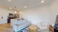 This thoughtfully designed single-floor residence offers a clean, contemporary aesthetic defined by bright white walls, light wood-look flooring, and recessed lighting throughout. The open-concept living area flows seamlessly into the kitchen, creating an inviting and functional shared space. The living room features generous seating space, accented by a geometric area rug and a gold metallic coffee table that adds a warm, modern touch.

The kitchen is a standout feature, equipped with a full suite of stainless steel appliances including a gas range, over-the-range microwave, French door refrigerator, and dishwasher. Crisp white upper cabinets contrast with rich dark lower cabinets, and the peninsula island with a white quartz countertop and undermount sink provides casual bar seating beneath elegant pendant lighting.

The unit includes two bedrooms. One bedroom is a well-lit, spacious room with a large window offering exterior views and ample floor space. The second bedroom features a built-in closet with organized shelving. Both rooms are finished with the same light flooring for a cohesive feel.

The bathroom is distinctively appointed with warm wood-look tile flooring and matching wall tile in the shower-tub combination, creating a striking contrast against the clean white fixtures and cabinetry. A walk-in closet with wire shelving is accessible from the bathroom area, adding practical storage.