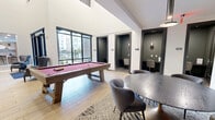 This thoughtfully designed single-floor amenity space offers a generous and versatile layout across a well-appointed floorplan. The main living area serves as the centerpiece, boasting soaring double-height ceilings, a sleek linear fireplace framed in rich wood and dark stone, built-in shelving niches with brass wall sconces, and bold large-scale artwork that commands attention. Plush seating in warm terracotta tones anchors the sophisticated lounge atmosphere. Light wood flooring flows throughout, unifying the space.

A second living and game area features floor-to-ceiling windows that flood the room with natural light and outdoor views, alongside a full-size billiards table and a large-screen TV. A casual dining nook with a channeled green velvet banquette and patterned tile flooring offers an intimate setting for smaller gatherings. A round conference table with upholstered seating provides a more formal meeting area.

The kitchen is outfitted with marble countertops, warm wood cabinetry, a bold green tile backsplash, patterned encaustic tile flooring, and globe pendant lighting — both functional and visually striking. Private office pods with built-in desks, moody green-painted walls, and individual pendant lighting offer quiet, focused workspaces. Two bathrooms complete this well-rounded amenity suite.
