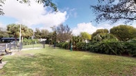 This outdoor space functions as a dedicated dog park, thoughtfully designed for pets and their owners. The grounds are enclosed by a sturdy chain-link fence that runs the perimeter, providing a secure area for animals to roam freely. The fenced enclosure features a gated entry point, ensuring safe access and containment.

The interior of the park is covered in well-maintained green grass, offering ample open space for recreation. Waste disposal stations are positioned within the park for convenience. A wooden bench is situated just outside the enclosure near the entry gate, providing a comfortable resting spot for visitors.

The surrounding landscape is lush with a variety of mature trees, including palms and broad-canopied shade trees, which frame the space and contribute to a verdant, tropical atmosphere. The natural canopy provides partial shade across portions of the grounds. The park enjoys open sky views, with natural light freely illuminating the area throughout the day.

Adjacent to the park, a paved pathway and parking area are visible, enhancing accessibility for visitors arriving by vehicle. The overall ambiance is open, airy, and well-suited for outdoor activity, reflecting a clean and community-oriented setting.