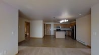 This single-floor space offers an open-concept layout that seamlessly connects the kitchen and living areas, creating a functional and cohesive environment. The kitchen is well-appointed with warm honey-toned wood cabinetry providing ample storage, a central island that serves as both a prep space and a casual gathering point, and a suite of stainless steel appliances including a refrigerator, range, and microwave. A chandelier-style light fixture overhead adds a touch of character to the kitchen area. The transition from the tile flooring in the kitchen to the carpeted living area subtly delineates the two zones while maintaining the open feel of the space. Neutral-toned walls and light-colored ceilings contribute to a bright and airy atmosphere throughout. A paneled interior door is visible, leading to additional rooms, including what appears to be a bathroom or bedroom corridor, suggesting further private living spaces beyond the main open area. The overall design is practical and welcoming, with a straightforward layout that maximizes usable space and flow.