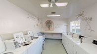Located in Encinitas, this single-story shared laundry facility offers a surprisingly welcoming atmosphere, distinguished by its bright, all-white interior and decorative botanical murals painted across the walls. The hand-painted artwork, depicting flowing vines, flowers, and a central arched motif showcasing a potted plant, lends the utilitarian space an unexpectedly warm and inviting character.

The room is well-equipped with a row of top-loading washing machines lining one wall, complemented by a corresponding bank of dryers along the opposite side. Ample folding and sorting space is provided by a long wall-mounted counter, ideal for organizing laundry efficiently. A garment hanging rack is also available for delicate items or freshly laundered clothing. White wicker seating is positioned near the folding area, offering a comfortable spot while waiting.

Illumination is provided by flush-mounted fluorescent ceiling panels and a ceiling fan, ensuring the space remains bright and well-ventilated. A window along one wall allows natural light to enter and provides views to the exterior. Near the entry door, a community bulletin board keeps residents informed. The light green flooring ties the clean, cohesive aesthetic together, making this laundry facility a functional yet pleasant shared amenity.