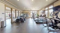 This fitness center is thoughtfully designed to support a wide range of workout routines across multiple distinct zones. The entry area sets the tone with polished aggregate flooring, a dark-stained wood shelving unit stocked with fresh towels, a decorative basketball hoop, and a seating bench — offering a functional and welcoming transition into the facility.

The main workout room is generously proportioned, featuring durable tile flooring and an abundance of natural light streaming in through large windows on multiple walls. A ceiling fan overhead aids in air circulation during intense sessions. The room is outfitted with a comprehensive selection of cardio equipment, including treadmills and stationary bikes, as well as strength training machines. Full-length mirrors line one wall, enhancing the sense of openness and allowing users to monitor their form. A wall-mounted flat-screen television adds an entertainment option for cardio workouts.

Adjacent to the main gym is a dedicated studio space, ideal for group fitness classes or individual flexibility and balance training. This room also benefits from large windows that flood the space with natural light, and features floor-to-ceiling mirrors, a ceiling fan, spinning bikes, and a variety of medicine balls and stability equipment. Together, these spaces create a comprehensive and inviting fitness facility.