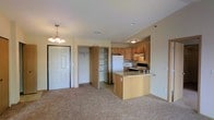This single-floor residential space offers a practical and comfortable open-concept layout that seamlessly connects the living area to the kitchen. The living area features neutral carpeting that adds warmth underfoot, and is illuminated by an elegant chandelier that serves as a decorative focal point near the entryway. The front entry door opens directly into this welcoming space, with a generously sized closet nearby providing convenient storage.

The kitchen is well-appointed with warm wood-toned cabinetry that extends to the upper walls, offering plentiful storage for cookware and pantry essentials. A peninsula with a built-in sink and countertop workspace creates a natural divide between the kitchen and living area while also providing additional seating or prep space. Appliances include a full-size refrigerator, a microwave, and a range with hood ventilation, making the kitchen fully functional and move-in ready. A second storage closet off the main living space adds further organizational versatility.

The overall ambiance of the space is bright and neutral, with light-toned walls and consistent flooring choices that give the unit a clean, cohesive feel. The layout is efficiently designed to maximize usability, making it a well-rounded residential space suitable for everyday living.