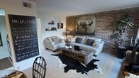 This single-floor space showcases a thoughtfully designed living area that masterfully combines rustic charm with modern sophistication. The centerpiece of the room is a striking exposed brick accent wall, which adds warmth and texture to the space and serves as the perfect backdrop for a large-scale equestrian artwork that commands attention. 

The living area is furnished with a plush, neutral-toned sofa and matching armchair, creating a comfortable and cohesive seating arrangement. A reclaimed wood coffee table anchors the space, complementing the rustic aesthetic while adding an organic, earthy element. Beneath the furniture, a bold black-and-white cowhide rug adds a striking contrast to the light-toned carpeting and ties the room's design elements together.

Wall-mounted wooden shelves display curated decorative accents, adding personality and character to the space. A vintage-style chalkboard sign near the entryway contributes to the eclectic, urban-industrial ambiance. The light-colored walls and carpeting brighten the interior, balancing the darker textures of the brick wall and wooden furnishings. Overall, the space exudes a warm, inviting atmosphere with a well-curated blend of natural materials and contemporary design sensibilities.