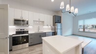 This thoughtfully designed one-bedroom, one-bathroom unit features a contemporary aesthetic throughout, unified by light wood-look flooring and crisp white walls. The open-concept kitchen and living area form the heart of the unit, anchored by a central kitchen island with a clean white countertop — ideal for dining or meal prep. The kitchen is fully equipped with stainless steel appliances, including a refrigerator, range, microwave, and dishwasher. Two-tone cabinetry pairs white upper cabinets with gray lower cabinets, complemented by a white subway tile backsplash for a polished, modern look. A striking multi-pendant chandelier defines the dining area, which flows seamlessly into a bright and airy living space. Large windows allow abundant natural light to fill the room, with views of a landscaped outdoor setting and access to a private balcony. The bedroom is a comfortable, light-filled retreat with a generous window and direct access to a walk-in closet outfitted with wire shelving for versatile storage. The bathroom features a wide gray vanity with a white countertop, a large mirror, and a full tub-shower combination, maintaining the unit's consistent modern style throughout.