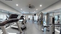 This fitness center is designed to support a comprehensive workout experience within a single-level layout. The space is furnished with a variety of cardio equipment, including treadmills and an elliptical machine, positioned along one side of the room. On the opposite side, a multi-station cable machine and an adjustable weight bench provide options for strength training. Full-length mirrors line the walls, enhancing the sense of openness and allowing users to monitor their form during exercise. The flooring consists of a durable, dark epoxy-style surface well-suited for high-traffic fitness use. Recessed lighting throughout the ceiling ensures the space is well-lit, while two ceiling fans contribute to air circulation and comfort during workouts. A window along one wall allows for natural light to enter the room. The neutral gray walls and clean finishes give the space a modern, functional aesthetic. Overall, the fitness center offers a practical and organized environment with a balanced selection of equipment suitable for both cardio and resistance training routines.