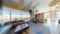 This elegantly designed single-floor communal space offers a seamless blend of contemporary architecture and refined interior design. The layout is expansive and open, anchored by soaring ceilings that amplify the sense of volume throughout. Floor-to-ceiling glass walls and oversized windows flood the interior with abundant natural light while framing sweeping panoramic views of the surrounding skyline and landscape. Dramatic blue botanical-print drapery panels add a bold artistic contrast against the light neutral palette of the walls and wide-plank hardwood flooring.

The lounge areas are thoughtfully arranged to encourage both relaxed conversation and quiet retreat. A central seating zone features a low-profile sectional in cool grey leather, accented by a geometric patterned area rug in blue, gold, and grey tones. Nearby, a cluster of mustard yellow and charcoal swivel chairs surrounds a round ottoman, creating a vibrant social hub. A pair of sleek shell chairs and a round accent table occupy a sun-drenched corner beneath the towering windows.

Sculptural gold honeycomb-style pendant lights serve as striking focal points throughout, complementing a decorative hexagonal wall installation in metallic tones. A built-in wood shelving unit with warm accent lighting anchors one wall, while elevator access and bar-height seating complete this polished, hospitality-inspired environment.