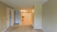 This single-floor space offers a straightforward, functional layout with a clear visual connection between the main living area and the kitchen beyond. The living area is finished with wall-to-wall carpeting in a neutral tone, complemented by soft beige walls and white baseboards that lend a clean, cohesive appearance throughout. A textured popcorn ceiling runs the length of the space, adding a traditional character overhead.

A wide opening leads from the carpeted living area into the kitchen, where the flooring transitions to a smooth, hardwood-style surface. The kitchen includes cabinetry and countertop space visible from the main room, making the overall layout feel open and connected. Flush-mount lighting illuminates the kitchen area effectively.

Off the living area, a walk-in closet provides generous storage space and features a distinctive brick accent wall, adding a subtle textural contrast to the otherwise smooth interior finishes. The space is further served by standard electrical outlets positioned conveniently along the walls.

Overall, the space presents a practical and well-maintained interior with a neutral palette, making it a versatile and move-in-ready option suitable for a variety of furnishing styles.