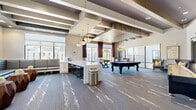 Located at 2007 Knoll Top Ln in Franklin, TN, this upscale amenity lounge delivers a well-appointed, single-floor gathering space designed for socializing and recreation. The open layout is anchored by a striking coffered ceiling with exposed wood-tone beams, recessed lighting, and a series of elegant chandelier fixtures that lend a warm, sophisticated ambiance throughout.

The game area is a central highlight, featuring a full-size pool table with a bold purple felt surface, a foosball table, and a shuffleboard table positioned along a textured accent wall with wall-mounted flat-screen televisions. Patterned carpet with an abstract white motif flows across the entire floor, adding visual depth and continuity.

A long white quartz-topped bar counter with barstool seating provides a functional gathering point, while a built-in banquette lounge area with padded bench seating and sculptural wooden side tables offers a comfortable spot for conversation. Large floor-to-ceiling windows and glass doors flood the space with natural light and frame views of the surrounding Franklin cityscape, enhanced by rich burgundy drapery panels.

Thoughtful decorative touches, including a guitar-motif wall art piece nodding to Tennessee's musical heritage, complete the character of this versatile and refined community space.