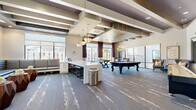 Located at 2007 Knoll Top Ln in Franklin, TN, this upscale amenity lounge delivers a well-appointed, single-floor gathering space designed for socializing and recreation. The open layout is anchored by a striking coffered ceiling with exposed wood-tone beams, recessed lighting, and a series of elegant chandelier fixtures that lend a warm, sophisticated ambiance throughout.

The game area is a central highlight, featuring a full-size pool table with a bold purple felt surface, a foosball table, and a shuffleboard table positioned along a textured accent wall with wall-mounted flat-screen televisions. Patterned carpet with an abstract white motif flows across the entire floor, adding visual depth and continuity.

A long white quartz-topped bar counter with barstool seating provides a functional gathering point, while a built-in banquette lounge area with padded bench seating and sculptural wooden side tables offers a comfortable spot for conversation. Large floor-to-ceiling windows and glass doors flood the space with natural light and frame views of the surrounding Franklin cityscape, enhanced by rich burgundy drapery panels.

Thoughtful decorative touches, including a guitar-motif wall art piece nodding to Tennessee's musical heritage, complete the character of this versatile and refined community space.