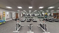 Located at 888 N Main St in Santa Ana, California, this single-floor fitness facility is designed to support a comprehensive workout experience. The space is anchored by a generously sized main exercise floor finished with durable, textured carpet tile and illuminated throughout by recessed LED panel lighting set within a drop ceiling — providing consistent, even light across all areas.

The cardio and strength zone is well-equipped with multiple treadmills and stationary spin bikes arranged in rows, all reflected in a full wall of mirrors that visually expands the room and allows users to monitor their form. Complementing the cardio machines, a freestanding dumbbell rack offers a range of free weights for strength training, while a colorful stability ball rack holds multiple sizes of exercise and medicine balls for functional training.

An open studio area adjacent to the main floor provides a versatile, unobstructed space ideal for group fitness classes, stretching, yoga, or personal training sessions. The studio also features mirrored walls and the same clean, neutral finishes found throughout the facility. Two interior doors lead to additional rooms, likely changing areas or storage. The overall design emphasizes functionality and cleanliness, with neutral gray walls, dark baseboards, and a no-frills aesthetic focused entirely on fitness utility.