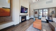 This contemporary single-floor amenity space is thoughtfully designed to blend style and functionality in a welcoming communal environment. The open-concept layout flows seamlessly from a well-appointed kitchen into a spacious lounge and gathering area, all unified by rich hardwood-style flooring throughout.

The kitchen is a standout feature, boasting dark wood cabinetry with sleek hardware, an expansive white quartz island with bar seating, and stainless steel appliances. A striking mosaic tile backsplash in muted tones of grey and white adds texture and depth, while open shelving and pendant lighting lend a modern, curated aesthetic.

The lounge area is anchored by a built-in fireplace topped with a wall-mounted television, framed by exposed brick accents and soaring ceilings that contribute to the space's airy, open feel. Clerestory windows allow natural light to flood in from above, complementing the large sliding glass doors that open directly to an outdoor pool deck. The panoramic views of the resort-style pool and surrounding community are a defining feature, creating a strong indoor-outdoor connection. A cozy seating nook positioned in front of the floor-to-ceiling grid windows makes for an ideal spot to take in the outdoor scenery. Overall, the space exudes a modern, upscale ambiance suited for both social gatherings and relaxed everyday use.