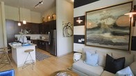 This single-floor space showcases a thoughtfully designed open-concept layout that seamlessly connects the kitchen and living area, creating a cohesive and functional environment. The kitchen is outfitted with dark espresso cabinetry, stainless steel appliances including a full-size refrigerator, and a light-toned countertop. A peninsula with bar-height seating adds both utility and a casual dining option. Warm gold pendant light fixtures hang above, complementing the rich cabinetry tones, while track lighting along the ceiling provides additional illumination. Light hardwood-style flooring flows throughout, lending a bright and airy feel to the space.

The living area makes a strong visual statement with a bold horizontal stripe accent wall in black and white, anchored by a large-scale, gold-framed landscape painting. A wall sconce adds a warm glow, enhancing the curated, gallery-like atmosphere. A neutral upholstered chair with a soft blue accent pillow contributes to the relaxed yet refined ambiance. Abstract artwork displayed between the two areas adds an artistic continuity. The overall design balances modern sophistication with inviting warmth.