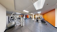 Located at 6358 Lancaster Avenue in Philadelphia, PA, this fitness center offers a generously sized, single-floor exercise room designed to accommodate a wide range of workout needs. The space features durable rubber tile flooring throughout, providing both safety and comfort during exercise. A drop ceiling with recessed panel lighting and skylights ensures the room is well-illuminated throughout.

The exercise room is organized into distinct functional zones. A dedicated cardio area houses equipment including a treadmill, recumbent bike, elliptical trainer, and rowing machine, with a wall-mounted flat-screen television positioned nearby for entertainment. The strength training section is well-appointed with a multi-station cable machine, free weights, kettlebells, medicine balls, and a weight bench, all complemented by large wall mirrors that enhance the sense of space and allow users to monitor their form.

Vibrant orange accent walls add energy to the environment, contrasting with the neutral cream-colored walls and white support columns throughout the room. The open floor plan allows for flexible use of the space, with ample room for stretching or functional fitness activities. The facility is accessible via a standard door with an emergency exit, ensuring convenience and safety for all users.