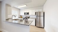 Located at 1201 Seminole Blvd in Largo, Florida, this single-floor residence offers a clean, modern aesthetic throughout its well-designed layout. The kitchen is a standout feature, showcasing white shaker-style cabinetry, granite countertops, and a full suite of stainless steel appliances including a refrigerator, electric range, microwave, and dishwasher. A peninsula counter provides additional workspace and opens toward the adjacent living and dining area, which is bathed in natural light from large windows fitted with white horizontal blinds. Light-toned wood-look flooring flows through the main living areas and hallways, lending a cohesive, contemporary feel. A bright, airy hallway with a textured ceiling connects the rooms, featuring a louvered closet door for convenient storage access. The bedrooms are generously sized and finished with plush carpeting, offering a comfortable retreat. Windows allow ample natural light into the sleeping quarters. The bathrooms are neatly appointed, featuring granite-topped vanities with white cabinetry, wood-look flooring, and a tub-shower combination. Throughout the residence, a neutral color palette of soft whites and warm grays creates a bright, welcoming atmosphere that is both functional and visually cohesive.