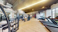This fitness center presents a thoughtfully designed workout space that blends industrial aesthetics with contemporary finishes. The exposed ceiling with visible ductwork and dark-painted structural elements contrasts with light hardwood-style flooring, creating a dynamic visual character throughout. Linear LED lighting runs along the ceiling, supplemented by recessed spotlights that ensure even illumination across the entire floor.

The space is organized into distinct functional zones. A cardio section lines the perimeter, featuring treadmills, stair climbers, rowing machines, and stationary bikes, all positioned to take advantage of the floor-to-ceiling steel-framed windows that frame lush outdoor greenery and palm trees. A dedicated strength training area anchors the center of the room, where a striking series of evenly spaced vertical wood-toned slat dividers creates a structural focal point while subtly defining workout zones without fully enclosing them.

A free weights station along a deep red accent wall offers a full dumbbell rack reflected by large mirrors, enhancing depth and utility. An attached restroom with a round mirror and sink provides added convenience. Wall-mounted televisions and vibrant artwork contribute to an energetic, motivating ambiance throughout the space.