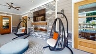 Located at 125 River Birch Grove Rd. in Asheville, NC, this single-level outdoor amenity space at Hawthorne at River Birch combines crafted architectural details with resort-style comfort. The exterior entrance is framed by stacked-stone columns, lap siding, and a warm wood-and-glass door, setting an inviting tone from the moment of arrival.

The centerpiece of the covered outdoor lounge is a dramatic floor-to-ceiling stacked-stone accent wall housing a linear gas fireplace topped with a rustic wood mantel, complemented by a wall-mounted television — ideal for year-round outdoor entertaining. Overhead, ceiling fans ensure comfortable airflow throughout the covered space.

The lounge area is furnished with hanging egg chairs and plush seating accented with bold orange throw pillows, all arranged over a patterned area rug. The space flows seamlessly to an open-air poolside terrace, where additional seating and dining options offer direct views of the swimming pool and surrounding lush greenery. Stone columns and black metal railings frame the terrace, reinforcing the cohesive design language throughout.

The interior fitness center, visible through the entrance door, completes the amenity suite. Together, these spaces deliver a well-appointed, community-focused environment that balances refined design with functional outdoor living in the heart of Asheville.