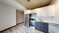 Located at 333 SW Harrison St in Portland, Oregon, this modern urban residence showcases a sleek industrial aesthetic throughout its single-floor layout. Exposed concrete ceilings lend an authentic urban character to the living spaces, complemented by light wood-look flooring and bold dark baseboards that create a striking visual contrast.

The kitchen is efficiently designed and outfitted with a full suite of stainless steel appliances, including a refrigerator, electric range, and over-the-range microwave. Two-tone cabinetry — white uppers paired with dark lower cabinets — adds contemporary style, while a stainless steel sink completes the functional workspace. A textured concrete accent wall adds visual depth to the kitchen area.

The living area benefits from generous floor-to-ceiling windows that invite abundant natural light and offer direct views of the surrounding Portland cityscape. Track lighting mounted to the concrete ceiling provides adjustable illumination throughout.

The bedroom features neutral carpeting, a ceiling fan, and a sliding door for privacy, maintaining the unit's clean, modern sensibility.

The bathroom is finished with light gray walls, wood-look tile flooring, and a full soaking tub with shower. A space-saving stacked washer and dryer unit is conveniently tucked into a dedicated in-unit laundry closet, adding practical functionality to this well-appointed urban residence.