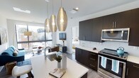 This single-floor space showcases a thoughtfully designed open-concept layout that seamlessly connects the kitchen, dining, and living areas. The kitchen is a standout feature, equipped with sleek dark cabinetry that contrasts beautifully against crisp white countertops and a subway tile backsplash. Stainless steel appliances, including a range and built-in microwave, add a polished, modern touch. A central island with an undermount sink serves as both a functional workspace and a casual gathering point.

Three distinctive woven pendant lights hang above the island, lending an artistic focal point to the space. Large windows flood the interior with natural light while offering views of the surrounding streetscape, enhancing the open, airy atmosphere. The living area is visible from the kitchen, creating a cohesive flow throughout the unit. Light-toned flooring runs continuously through the space, reinforcing the clean, contemporary aesthetic. A glimpse into the bedroom reveals bright white walls and natural light, consistent with the overall modern design sensibility found throughout this well-appointed space.