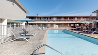 This property is built around a central courtyard that serves as the focal point of the outdoor amenities. The architecture follows a classic mid-century motel style, with two-story building wings wrapping around the pool area, featuring exterior walkways, metal railings, and individual room entrances accessible from open-air corridors. A staircase connects the upper and lower levels, and desert-friendly landscaping with rock ground cover and spiky plants accents the courtyard perimeter.

The outdoor pool is generously sized with clear, blue-tinted water and stainless steel entry rails. The surrounding deck features decorative stamped concrete in a stone-like pattern, providing ample space for sunbathing and relaxation. White lounge chairs and teal accent furniture are arranged throughout the pool deck, complemented by teal market umbrellas that provide shade during sunny days.

A standout feature of the courtyard is a stylish canopied daybed with a teal patterned frame and plush white cushions, positioned on the stamped concrete area. The open-air design of the space allows for abundant natural light and a bright, resort-like atmosphere under clear skies.