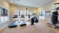This single-floor fitness center is thoughtfully designed to support a comprehensive workout experience. The space is organized into distinct functional zones, each tailored to a specific type of training. The cardio area features multiple treadmills, an elliptical, and a stair climber, all positioned along a wall lined with large windows that invite natural light and provide outdoor views, creating an energizing atmosphere for exercise. A ceiling fan overhead aids in air circulation throughout this section.

The strength training zone is anchored by a Life Fitness Synergy 360 multi-functional cable and resistance station, complemented by a hanging punching bag and a set of kettlebells, offering a versatile range of functional fitness options. A full rack of dumbbells and an adjustable flat bench provide additional free-weight training capabilities.

Floor-to-ceiling mirrors span one wall, visually expanding the space and allowing users to monitor their form during workouts. Informational charts and fitness rule signage are displayed throughout, adding a professional, facility-grade character to the environment.

The flooring consists of durable carpet tiles throughout, providing traction and comfort underfoot. A dedicated entry alcove includes a stainless steel dual water fountain with a bottle-filling station, and a glass-paned French door leads to an adjacent room, rounding out this well-appointed fitness facility.