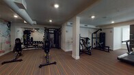 Located at 6150 Canoga Ave in the Woodland Hills neighborhood of Los Angeles, this fitness center is a thoughtfully designed, single-floor facility offering a comprehensive workout environment. The space features a striking striped hardwood floor with orange accent lines that runs throughout, providing a cohesive and professional aesthetic.

The strength training area is well-appointed with a variety of cable machines, multi-functional weight systems, and adjustable benches, accommodating both compound and isolation exercises. A dedicated free weights section includes a full dumbbell rack, reflected by wall-mounted mirrors that enhance the sense of space and allow users to monitor their form.

A separate cardio zone features a curved wall of large windows fitted with roller shades, allowing natural light to fill the area while providing privacy. This section houses a row of Matrix-brand recumbent bikes and elliptical trainers, creating a bright and motivating environment for cardiovascular workouts. Additional cardio equipment, including a treadmill, is positioned throughout the floor.

Recessed ceiling lighting illuminates the entire space evenly, complemented by exposed ductwork in select sections that adds an industrial touch. Structural columns are cleanly finished in white, integrating seamlessly into the overall design. Sanitation stations are conveniently placed throughout the facility, and clearly marked emergency exits ensure safety. The overall ambiance is clean, modern, and purpose-built for fitness.
