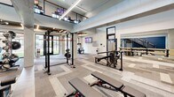 Located at 185 Marquis Way in Freeport, Florida, this two-floor fitness facility offers a well-appointed and thoughtfully designed space for comprehensive exercise. The main floor is the larger of the two levels, featuring an open, airy layout with striking double-height ceilings that create a sense of expansive volume. A slatted wood ceiling detail near the entrance adds warmth and architectural interest, complemented by large glass doors that flood the space with natural light and provide direct access to an outdoor green area. Patterned carpet tiles in a chevron-inspired design cover the floors throughout, lending a modern, polished aesthetic. The main exercise room is equipped with strength training stations, a power rack, free weights, and benches, alongside wall-mounted televisions for entertainment. A bold navy accent wall adds a dynamic visual anchor to the space. The upper floor, accessed via a staircase with a sleek wood-and-cable railing, offers a more intimate exercise area dedicated to cardio equipment, including ellipticals, stationary bikes, and a rowing machine. Wall-mounted televisions and windows overlooking the surrounding community keep this level bright and engaging. Together, the two floors provide a well-rounded, modern fitness environment suited for a full range of workout routines.