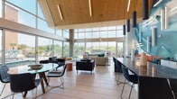 Located at 6001 S Congress Ave in Austin, Texas, this single-floor clubhouse is a standout example of contemporary communal design. The space is defined by its dramatic vaulted ceiling finished in warm natural wood planking, which creates an inviting architectural centerpiece and amplifies the sense of openness throughout. Expansive floor-to-ceiling glass walls wrap around the perimeter, flooding the interior with abundant natural light and offering unobstructed views of the surrounding outdoor amenity areas, including a pool and landscaped grounds.

The main lounge area is generously proportioned, featuring multiple distinct seating arrangements that cater to both social gatherings and quiet relaxation. Furnishings include a large sectional sofa with teal accent pillows, sleek black leather sofas, red accent chairs, and a variety of coffee and side tables in glass and mixed materials. Large-format light tile flooring runs throughout, enhancing the bright, airy atmosphere.

A striking teal and white abstract mural serves as a bold focal point on one interior wall, adding an artistic dimension to the space. A kitchenette area with a dark marble island, white cabinetry, and a sink provides functional convenience. Pendant lighting and linear LED fixtures complement the natural light. Glass partitions and sliding doors connect the interior seamlessly to adjacent outdoor spaces, reinforcing the open, community-oriented character of this well-appointed facility.