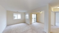 Located at 1 Greenview Circle in West Sayville, NY, this single-floor unit at Greenview West offers a bright and open layout designed for comfortable living. The main living area is generously proportioned, featuring plush wall-to-wall carpeting in a warm neutral tone that extends throughout the space. Cream-colored walls and smooth ceilings create a clean, airy atmosphere, while multiple windows along the exterior walls allow abundant natural light to fill every corner of the room. A ceiling fan with a light fixture is positioned in the adjoining area, providing both air circulation and additional illumination. A sliding glass door connects the interior living space directly to an outdoor area, offering convenient access and views of the surrounding greenery. The open-concept flow between the living and adjacent room provides a seamless and functional layout. The kitchen area, visible from above, features light cabinetry and tiled flooring, providing a practical workspace. The unit's single-floor design ensures easy accessibility throughout, with a straightforward and efficient floor plan. Overall, the space presents a light-filled, well-maintained interior with a neutral palette that offers a versatile foundation for a variety of living arrangements.