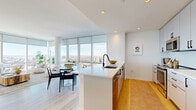 Located in Jersey City's vibrant downtown, this high-rise residence at 180 Morgan Street offers four bedrooms, three bathrooms, and a thoughtfully designed open layout bathed in natural light. The living room is a standout centerpiece, featuring dramatic floor-to-ceiling windows that frame sweeping panoramic views of the New York City skyline and surrounding cityscape, creating a bright and airy atmosphere throughout the day.

The kitchen is sleek and modern, appointed with white cabinetry, stainless steel appliances including a full-size refrigerator, range, and microwave, along with a central island with a waterfall-edge countertop and an integrated sink. Light hardwood flooring runs continuously through the main living areas and hallway, lending warmth to the clean, contemporary aesthetic.

The four bedrooms are well-proportioned and filled with natural light; the primary bedroom benefits from expansive city views and generous closet space with double built-in storage. The three bathrooms are finished with crisp white tile, modern vanities, and black hardware accents for a refined, contemporary look.

A long central hallway connects all rooms efficiently and includes in-unit stacked laundry. The overall design palette — white walls, light wood floors, and minimalist finishes — creates a cohesive, modern living environment with striking urban views as a constant backdrop.