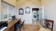 This single-floor business center offers a refined and functional environment designed to meet professional needs in a welcoming setting. The room is anchored by a warm, dark-wood desk equipped with a desktop computer and multifunction printer, providing all the essentials for productivity. A standout architectural feature is the large arched window fitted with classic plantation shutters, which floods the space with abundant natural light while framing views of the surrounding landscape. The vaulted ceiling adds a sense of openness and grandeur to the room. Decorative touches throughout lend a sophisticated ambiance — ornate framed artwork, an intricate metal wall medallion, a decorative wall clock, and an elegantly framed mirror work together to create a polished aesthetic. Comfortable seating with upholstered cushions is available for guests, complemented by a wrought-iron plant stand adorned with fresh florals. Patterned carpeting and tile flooring add texture and warmth to the space. An adjacent fitness room, visible through glass-paneled doors, extends the utility of this amenity area. Overall, the business center strikes a balance between practicality and tasteful décor, making it a welcoming and fully equipped space for work.