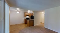 This single-story space offers an open-concept layout that seamlessly connects the living, dining, and kitchen areas. The kitchen is equipped with medium-toned wood cabinetry, dark granite-style countertops, and black appliances, including a refrigerator and range. A peninsula with a breakfast bar extends into the living space, providing both additional prep space and casual seating. Track lighting above the kitchen illuminates the work areas effectively.

The living area features neutral carpet flooring, which contrasts with the hardwood-style flooring found in the kitchen and hallway. A ceiling fan with an integrated light fixture is centrally positioned in the living space, offering both air circulation and ambient lighting. The textured popcorn ceiling is consistent throughout the unit, adding a classic residential feel.

A hallway visible from the main living area leads to additional rooms, with a bi-fold closet door suggesting convenient storage. The overall color palette is neutral, with warm beige walls and light-toned trim, creating a clean and functional living environment suitable for everyday use.