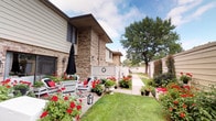 This single-story property combines attractive exterior design with practical shared amenities. The building's exterior showcases classic brick construction complemented by vinyl siding, giving it a timeless, well-maintained appearance. A standout feature of the property is its thoughtfully curated outdoor space, where a private patio area runs alongside the building. The patio is adorned with abundant red geraniums and flowering plants arranged in decorative urns and containers, creating a vibrant, garden-like atmosphere. White wrought-iron patio furniture, including a dining table and chairs with red accent cushions, provides a comfortable outdoor seating area, while a black market umbrella offers shade. A concrete walkway borders a neatly manicured lawn, and a privacy fence with a decorative clock accent separates the space. Mature trees in the background add to the serene setting.

The building also includes a shared laundry facility, equipped with multiple LG commercial washers and dryers, a utility sink, and posted operating instructions. The laundry room features tile flooring, neutral-toned walls, and a textured ceiling. Access to this utility space is provided through a glass-paneled entry vestibule with a mat floor and a wall-mounted intercom or vent unit.