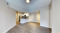 This single-floor space showcases an open-concept layout that seamlessly connects the living area and kitchen, creating a spacious and functional environment. The flooring throughout consists of warm-toned hardwood-style planks, lending a cohesive and contemporary feel to the space.

The kitchen is well-appointed with white cabinetry, stainless steel appliances including a refrigerator and microwave, and a tasteful tile backsplash that adds texture and visual interest. A peninsula with a light-coloured countertop extends into the living space, providing a casual breakfast bar area ideal for dining or entertaining. Track lighting installed above the kitchen ensures ample task illumination.

The living area is generously proportioned, offering flexible arrangement possibilities. Neutral grey-toned walls create a clean, versatile backdrop suitable for a variety of décor styles. A ceiling light fixture provides central illumination for the living space. Crisp white baseboards frame the room neatly, complementing the overall modern aesthetic. A hallway visible in the background suggests additional rooms or storage beyond the main living and kitchen area.