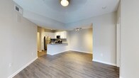 This single-floor space showcases an open-concept layout that seamlessly connects the living area and kitchen, creating a spacious and functional environment. The flooring throughout consists of warm-toned hardwood-style planks, lending a cohesive and contemporary feel to the space.

The kitchen is well-appointed with white cabinetry, stainless steel appliances including a refrigerator and microwave, and a tasteful tile backsplash that adds texture and visual interest. A peninsula with a light-coloured countertop extends into the living space, providing a casual breakfast bar area ideal for dining or entertaining. Track lighting installed above the kitchen ensures ample task illumination.

The living area is generously proportioned, offering flexible arrangement possibilities. Neutral grey-toned walls create a clean, versatile backdrop suitable for a variety of décor styles. A ceiling light fixture provides central illumination for the living space. Crisp white baseboards frame the room neatly, complementing the overall modern aesthetic. A hallway visible in the background suggests additional rooms or storage beyond the main living and kitchen area.