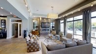 This single-story clubhouse delivers a sophisticated and welcoming atmosphere designed for comfort and socializing. The main lounge area is the centerpiece of the space, showcasing a refined interior design with a neutral palette accented by gold and warm metallic tones throughout. A striking stone-clad fireplace serves as the focal point of the room, flanked by decorative cabinetry and topped with a large flat-screen television, making it ideal for both relaxed gatherings and entertainment.

The seating area is thoughtfully arranged with plush sofas and accent chairs, including patterned upholstery that adds visual interest to the sophisticated décor. A glass-topped coffee table and a modern chandelier with cascading pendants contribute to the upscale ambiance. Track lighting along the ceiling provides adjustable illumination throughout the space.

Expansive floor-to-ceiling windows with elegant drapery line one wall, flooding the interior with natural light while offering direct views of the outdoor pool and lounge area beyond. A large decorative mirror enhances the sense of openness and depth. The tile flooring is durable yet refined, complementing the overall design aesthetic. Together, these elements create a polished, resort-style clubhouse environment built for community and leisure.