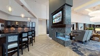 This single-floor lobby space showcases a contemporary design aesthetic defined by clean lines, high ceilings, and an open, airy layout. The centerpiece of the room is a striking double-sided fireplace clad in textured stacked-stone tile, rising dramatically toward the ceiling. A large flat-screen TV is mounted directly above the fireplace, making it a commanding focal point visible from multiple areas of the space.

The flooring features a herringbone-patterned light wood design that adds visual interest and warmth throughout. To one side, a bar or refreshment station offers high-top seating with dark wood chairs, complemented by pendant globe lighting and glass-front cabinetry that adds a refined, lounge-like quality. The opposite side transitions into a comfortable seating lounge area with upholstered sofas and a patterned area rug in deep blues.

Soaring vaulted ceilings enhance the sense of openness, while the neutral white and gray color palette, accented by dark stone and wood tones, creates a sophisticated yet inviting atmosphere well-suited for a hospitality or communal gathering environment.