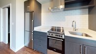 This single-floor unit offers a thoughtfully designed layout with a cohesive modern aesthetic throughout. Warm hardwood-style flooring runs continuously across the living areas and bedrooms, providing a consistent and inviting foundation. The neutral gray wall tones complement the flooring and create a calm, versatile backdrop throughout the space.

The kitchen is a standout feature, equipped with dark wood cabinetry, white quartz-style countertops, and a full suite of stainless steel appliances including a refrigerator, electric range with overhead ventilation hood, and built-in microwave. A classic white subway tile backsplash adds a clean, contemporary touch. A large window or sliding door with vertical blinds allows natural light to fill the kitchen and adjacent living area.

The bathroom is well-appointed with white subway tile surrounding a full soaking tub and shower combination, wood-style flooring, and a vanity with dark cabinetry. A dedicated laundry area houses a stacked washer and dryer unit, offering practical convenience within the unit. The bedrooms are generously sized, featuring neutral walls, hardwood-style floors, and access to natural light through windows with blinds.
