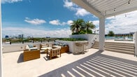 Located at 2585 Spruce Ave in West Palm Beach, FL, this rooftop amenity deck offers a resort-caliber outdoor experience with a clean, modern aesthetic. The centerpiece is a generously sized swimming pool finished with teal tile accents, surrounded by an expansive sun deck lined with rows of turquoise lounge chairs and sleek white sun loungers — providing ample space for relaxation under the Florida sky.

The terrace extends well beyond the pool area, offering multiple distinct zones for outdoor enjoyment. A shaded pergola structure with a slatted louvered roof covers a stylish outdoor living area furnished with teak-framed seating, plush cushioned sofas, and dining tables, ideal for entertaining or casual gatherings. String lights add a warm ambiance to the covered space, while an outdoor kitchen with grilling stations serves as a functional focal point.

A glass-enclosed interior lounge space adjoins the rooftop terrace, featuring floor-to-ceiling windows that frame the cityscape and flood the interior with natural light. High-top seating and casual bar-style tables complete this transitional indoor-outdoor area.

Throughout the deck, panoramic views of the West Palm Beach skyline, lush treetops, and the wide South Florida horizon create a striking backdrop. The overall design blends contemporary white architecture with vibrant coastal accents, delivering a polished, resort-like atmosphere.