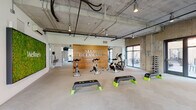 Located on W 2nd Street in Kansas City, Missouri, the Second + Delaware fitness center is a well-appointed, single-floor wellness facility designed with a contemporary industrial aesthetic. Exposed concrete ceilings with visible ductwork and piping contrast with clean white walls and warm wood accents, creating a polished yet raw atmosphere throughout the space.

The facility is organized into distinct functional zones. A group exercise studio anchors one end of the space, featuring a bold wood-paneled feature wall bearing the Second + Delaware branding, a living moss wall labeled "Wellness," step platforms, stationary spin bikes, and an air bike. Large glass doors in this area open directly to an outdoor patio, allowing ample natural light and fresh air to flow in.

A dedicated cardio zone is positioned near floor-to-ceiling windows overlooking the street, housing treadmills, elliptical trainers, and rowing machines, all bathed in natural light with mounted screens for entertainment. Adjacent to this is a strength training area equipped with a power rack, free weights, kettlebells, a balance trainer, and open floor space for functional movement.

Warm wood cabinetry and shelving provide practical storage, while herringbone-patterned flooring ties the zones together cohesively. The overall ambiance is energetic, modern, and inviting.