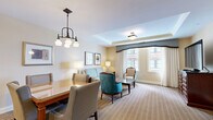 This elegantly designed one-bedroom suite offers a thoughtfully organized single-floor layout that balances comfort and functionality. The open-concept living and dining area is anchored by plush carpeting, a tray ceiling with crown molding, and large windows dressed in floor-length drapes that invite generous natural light. The living space features a classic seating arrangement with a light blue sofa, teal accent chairs, and a coffee table, creating a refined yet inviting atmosphere. Adjacent to the living area, the dining space accommodates a full-sized table with upholstered seating beneath a statement chandelier. The kitchen is outfitted with warm cherry wood cabinetry, granite countertops, stainless steel appliances including a full-size refrigerator, and pendant lighting along the breakfast bar. The spacious primary bedroom includes large windows with decorative valances, a dedicated workspace with seating, a dark wood media console with a flat-screen TV, and ample natural light. Five closets throughout the suite provide exceptional storage, including a walk-in closet with full-length hanging space and a luggage rack. The bathroom is finished with travertine tile, a soaking tub, a glass-enclosed shower, and a dark granite vanity with cherry wood cabinetry.