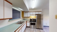 This two-floor space offers a practical and straightforward layout designed for comfortable everyday living. The kitchen is a functional highlight, featuring stainless steel appliances including a gas range and a full-size refrigerator. White cabinetry with warm wood trim accents lines the walls, providing ample storage, while light-toned countertops and a double-basin sink add to the kitchen's utility. Light wood-style flooring runs throughout the kitchen, and recessed fluorescent lighting ensures a well-lit workspace. A pass-through window connects the kitchen to the adjacent living area, which is carpeted in a neutral gray tone and offers an open, versatile layout.

The bedrooms are carpeted and finished with neutral walls, each featuring windows that bring in natural light and views of the surrounding neighborhood. The rooms are generously proportioned, offering flexibility for various furniture arrangements. The bathroom includes a standard toilet and a full tub and shower combination, finished in clean white tones with wood-style flooring. Throughout the space, dark baseboards provide a subtle contrast to the light-colored walls, contributing to a cohesive and understated aesthetic across both floors.