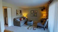 This single-floor space centers around a living room that exudes a warm and inviting ambiance, achieved through carefully layered lighting and coordinated furnishings. The room features soft, wall-to-wall carpeting that adds comfort underfoot and contributes to the overall coziness of the space. Neutral gray walls serve as a versatile backdrop, complemented by warm-toned lamp lighting that creates an intimate atmosphere throughout.

The seating area is anchored by a substantial gray sofa dressed with an eclectic mix of throw pillows in bold red, black-and-white geometric, and floral patterns, adding visual interest and personality. A matching gray ottoman serves as a functional centerpiece atop a patterned area rug featuring swirling earth tones of cream, burgundy, and tan. A gray accent chair provides additional seating to one side, while a dark wood side table and lamp complete the arrangement.

The walls are decorated with a canvas art piece featuring a warm, textured mosaic, and a grouping of four framed decorative mirrors with geometric metallic designs. Spherical woven pendant accessories in the corner add an organic, artisanal touch, rounding out the room's eclectic yet cohesive aesthetic.