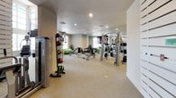 This contemporary fitness center is designed to offer a comprehensive workout experience within a bright, open, and thoughtfully organized space. The facility is anchored by soaring double-height ceilings in the main exercise area, with expansive floor-to-ceiling windows that flood the space with natural light and provide views of the surrounding exterior. Neutral-toned carpet flooring runs throughout, complementing the clean white walls and creating a welcoming, professional atmosphere.

The main floor is generously proportioned, accommodating a wide selection of commercial-grade strength machines, including Cybex cable systems and multi-use weight stations, along with free weights, medicine ball racks, and adjustable benches. A dedicated mirrored section enhances the sense of depth and functionality in the strength training zone. Cardio equipment, including a rowing machine and a stair climber, is also available.

A striking spiral staircase with sleek black metal railings and warm wood treads serves as both a functional and architectural centerpiece of the space. White-framed glass-panel doors provide access to distinct zones within the facility, adding structure without sacrificing openness. Recessed lighting throughout ensures consistent illumination across all workout areas, making this fitness center a polished and fully equipped destination for training.