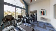 This fitness center is designed to offer a comprehensive and motivating workout environment across a single level. The space is divided into two connected zones — a cardio area and a strength training area — each thoughtfully appointed with modern equipment and design details.

The cardio zone is bathed in natural light, courtesy of expansive floor-to-ceiling windows framed by a dramatic arched transom. These windows frame views of lush outdoor surroundings, including palm trees and what appears to be a resort-style setting, lending an energizing backdrop to every workout. Soaring vaulted ceilings with ceiling fans enhance the sense of openness and airflow throughout this section. Treadmills and elliptical cross-trainers are arranged to take full advantage of the outdoor views.

The strength training area transitions seamlessly from the cardio zone and features a variety of cable machines, weight benches, and multi-functional strength equipment. A bold geometric accent wall in teal and gold adds a striking visual element, while ceiling fans maintain a comfortable atmosphere. Motivational artwork, including a large running-themed photo print, adds to the energetic ambiance.

Throughout both zones, the flooring consists of patterned modular carpet tiles in cool blue and gray tones, complementing the contemporary color palette of teal, white, and gray that defines the overall aesthetic of the space.