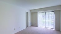This single-floor space offers a clean, functional layout designed for comfortable everyday living. The main living area is a standout feature, characterized by its bright and airy atmosphere, thanks largely to expansive floor-to-ceiling sliding glass doors dressed with vertical blinds. These doors flood the room with natural light while providing direct views of the lush greenery outside, creating a seamless connection between the interior and the outdoors. The neutral, white walls and light-toned carpeted flooring lend a sense of openness and calm throughout the space. A built-in wall vent ensures efficient climate control, contributing to the room's practical design. A doorway off the main living area provides access to additional rooms within the unit, maintaining a straightforward and efficient floor plan. The overall aesthetic is minimal and move-in ready, offering a blank canvas well-suited for a variety of interior styles and furnishing arrangements. The abundance of natural light, combined with the soft carpeting and clean wall surfaces, gives the space a welcoming and versatile character.