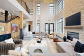 Located at 380 Crown Dr in Emerson, Georgia, this expansive single-floor space makes an immediate impression with its dramatic double-height ceilings and towering floor-to-ceiling grid windows that flood the interior with abundant natural light. The living room serves as the centerpiece, anchored by a striking stacked-stone fireplace and flanked by an arrangement of sophisticated seating areas that invite both relaxed gathering and conversation.

Architectural details are thoughtfully layered throughout. Structural columns clad in wood and dark metal add visual depth, while exposed wood ceiling beams lend warmth and texture across different zones of the space. Large-format slate-toned tile flooring transitions into rich wide-plank hardwood, defining distinct areas within the open layout. Arched wall niches add a refined Mediterranean-inspired touch, complementing the overall contemporary-rustic aesthetic.

A mezzanine-level overlook with metal railings is visible from the main living area, adding vertical drama and a sense of grandeur. An adjacent dining and lounge area features dark-framed glass doors opening to the outdoors, paired with rustic wood beam ceiling details and pendant lighting. Throughout, the design balances industrial elements—steel-framed windows, dark metal accents—with warm natural materials, creating a cohesive and inviting ambiance ideal for both everyday living and entertaining.